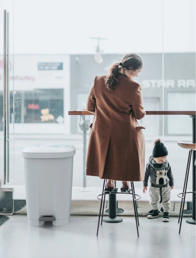 Mãe sentada a uma mesa com criança ao lado do Balde do Lixo Profissional com Pedal 50L em um ambiente moderno.