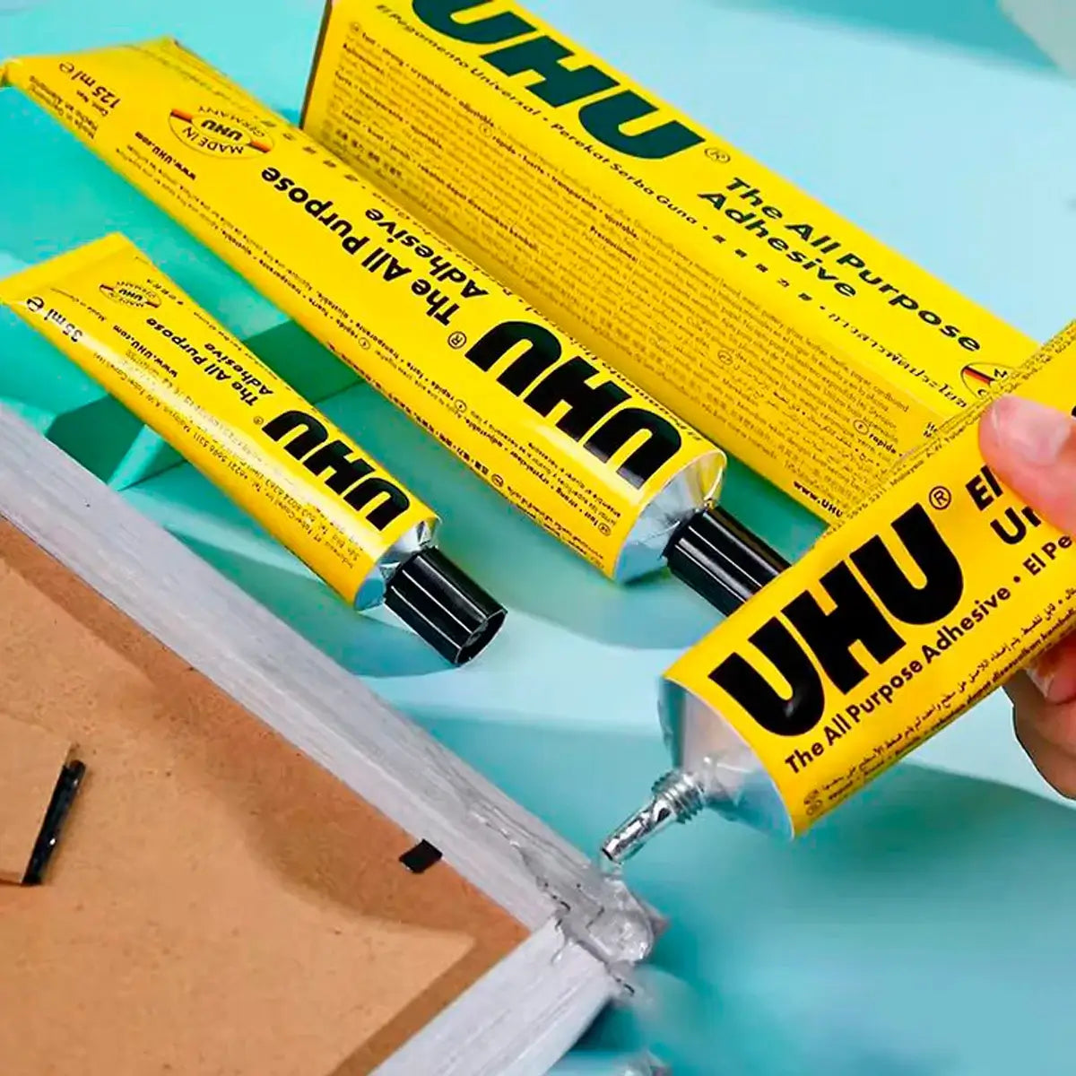Cola Líquida Universal Bisnaga 125ml UHU e outros adesivos da UHU em uso sobre superfície de trabalho.